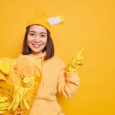 Pleased Asian Woman Does Laundry Home Wears Hat With Stuck Toilet Brush Hoodie Rubber Gloves Points Away Blank Copy Space Isolated Yellow Background Shows Product Cleaning
